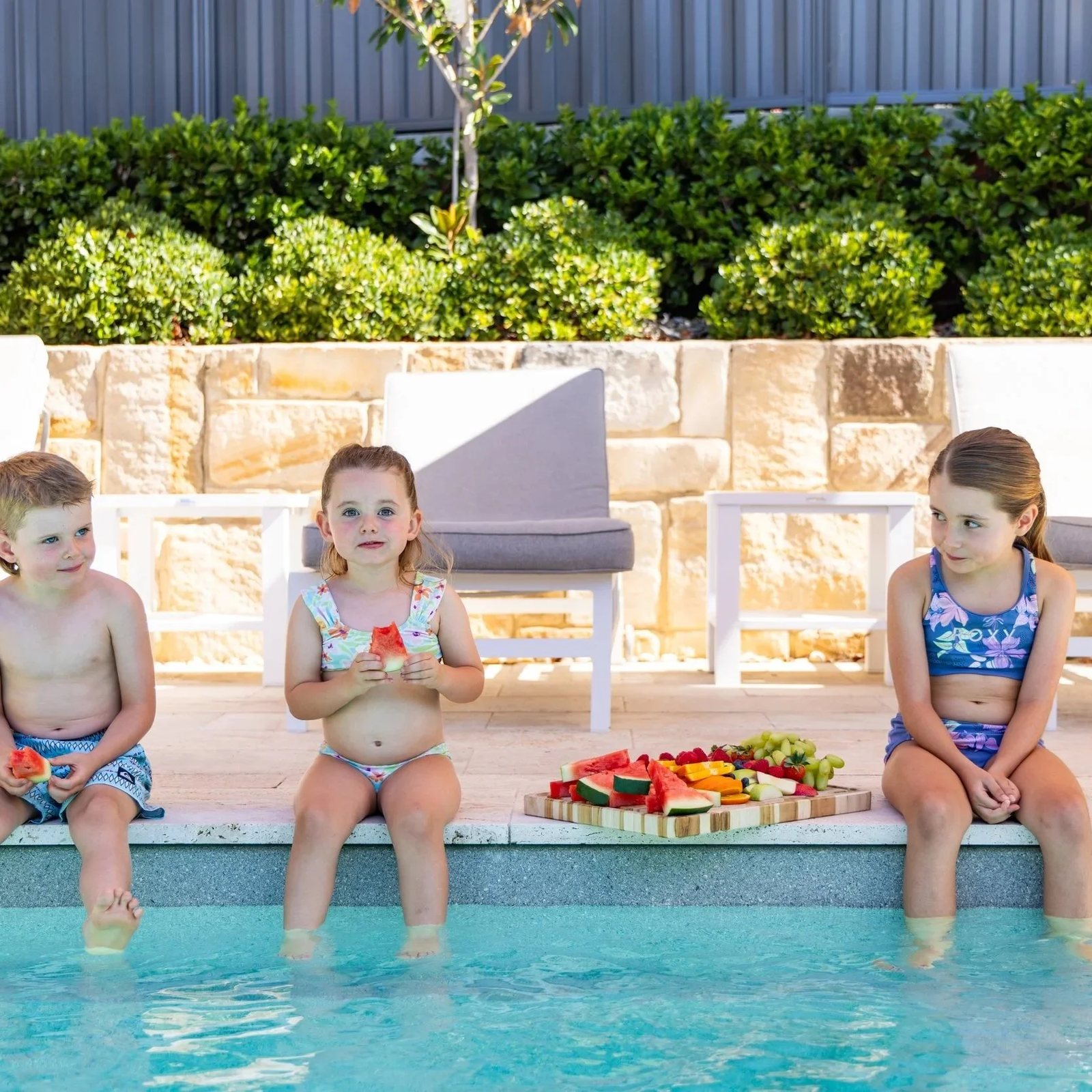 Three children sitting on the edge of a pool with a fruit platter behind them