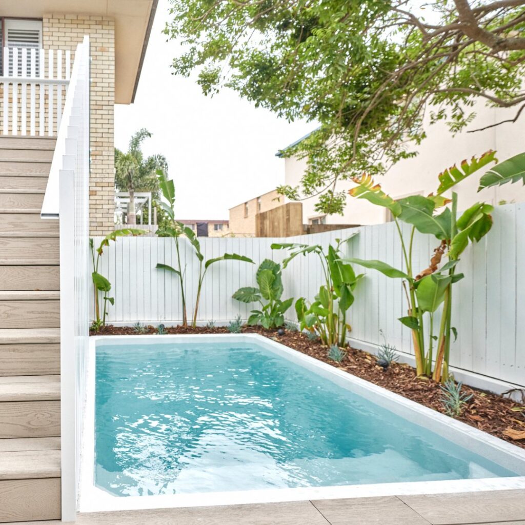 Compact backyard pool beside a white fence with tropical planting